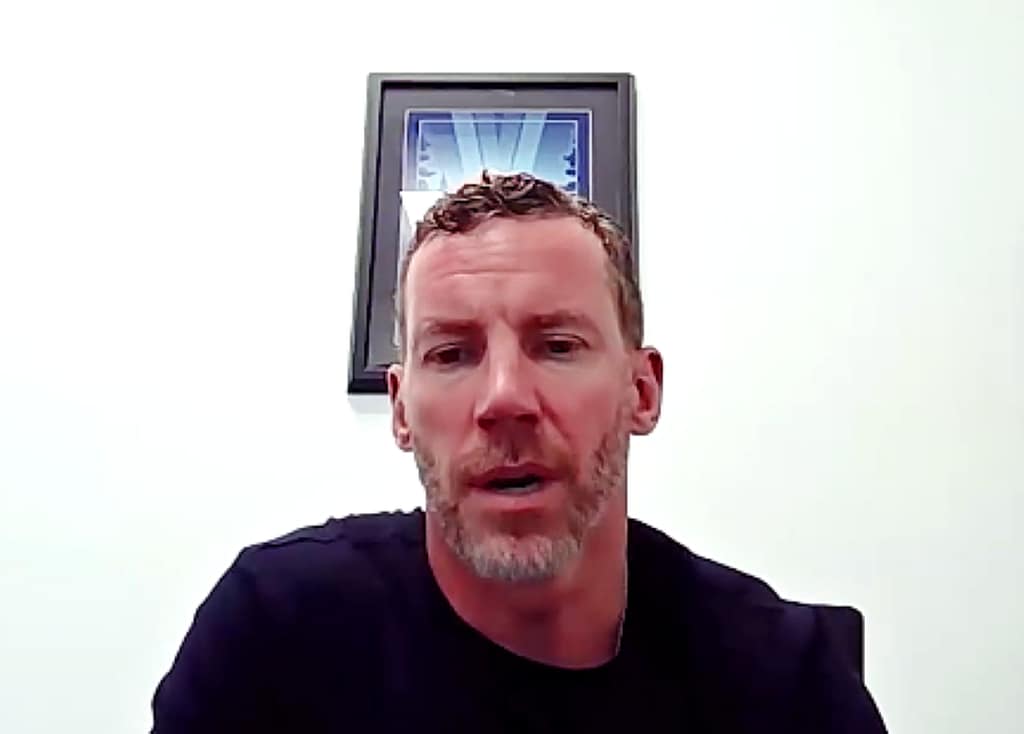 A man with short, tousled hair and a beard speaks in front of a white wall, displaying a framed picture in the background.