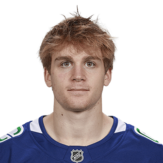 Headshot of a young male hockey player wearing a team jersey, with short, tousled hair and an expressionless face against a gray background.