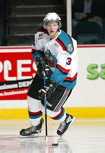 A hockey player wearing a white jersey with red and blue accents glides on the ice, holding a hockey stick and looking towards the puck.