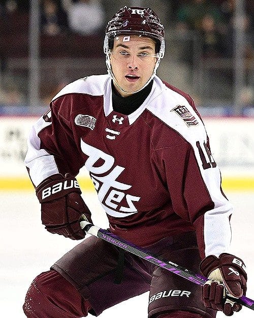 A male ice hockey player in a maroon and white uniform, focused and poised on the ice with his stick ready.