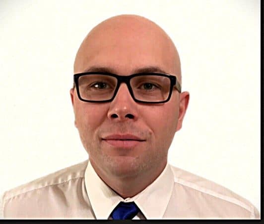 A man with a bald head and glasses, wearing a white shirt and a blue tie, poses against a plain white background.