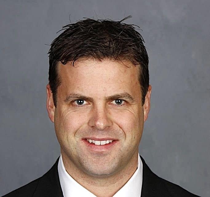 A smiling man with short dark hair, wearing a black suit and white shirt, against a plain gray background.